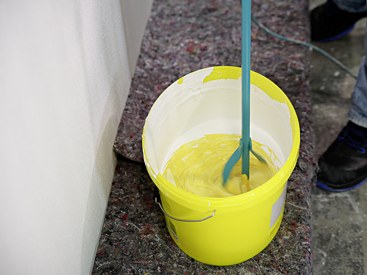 Nahaufnahme in einen gelben Produkteimer, in dem gelbe Farbe mit dem Rollputz mit einem Rührgerät vermischt wird.